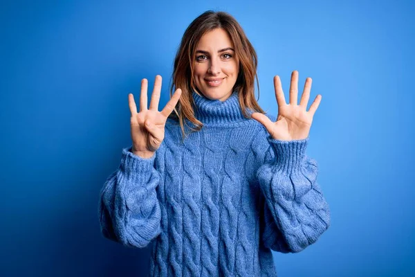 파란색 배경에 터틀넥 스웨터를 자신감 행복하게 웃으면서 손가락으로 가리키는 아름다운 — 스톡 사진