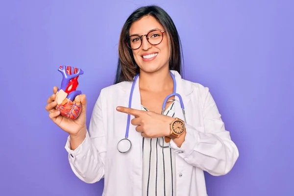Young doctor cardiology specialist woman holding medical heart over pruple background very happy pointing with hand and finger