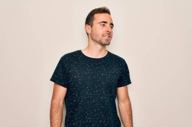 Mavi gözlü, sıradan tişörtlü, beyaz arka planda duran, yüzünde gülümseme, doğal bir ifade olan genç yakışıklı bir adam. Kendinden emin gülen.