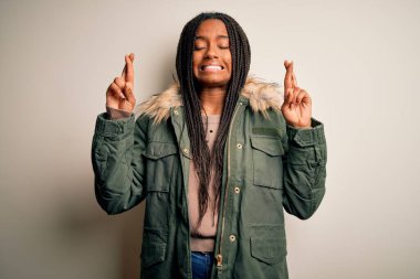 Genç Afro-Amerikalı kadın izole edilmiş arka planda kışlık parka ceket giyiyor umut ve gözler kapalı bir şekilde gülümseyerek el işareti yapıyor. Şans ve batıl inanç..