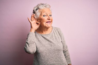 Üst düzey güzel bir kadın, günlük tişört giyip pembe arka planda dikiliyor. Kulaktan kulağa gülümsüyor, dedikodu ve dedikoduları dinliyor. Sağırlık kavramı.