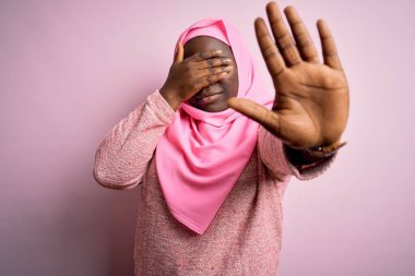 Genç Afro-Amerikan artı cüsseli kadın izole edilmiş pembe arka plan üzerine Müslüman başörtüsü takıyor gözleri ellerle örtüyor ve üzgün ve korku ifadesiyle dur hareketi yapıyor. Utanç verici ve olumsuz bir kavram..
