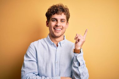 Sarışın, kıvırcık saçlı, sarı arka planda çizgili gömlek giyen, yüzünde kocaman bir gülümseme olan, eli ve parmağı kameraya bakan genç, yakışıklı bir adam..