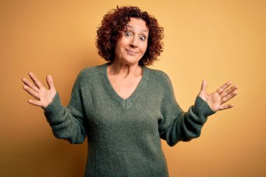 Orta yaşlı güzel kıvırcık saçlı kadın izole edilmiş sarı arka planda sıradan bir kazak giyiyor ve elleri ve kolları havada kafası karışmış bir ifade takınıyor. Şüphe kavramı.