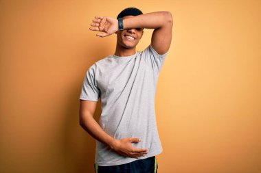 Yakışıklı Afro-Amerikan sporcu sarı arka planda spor giysisi giyiyor gözleri kollarıyla kapatıyor neşeli ve komik gülümsüyor. Kör kavram.