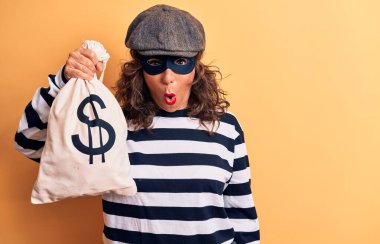 Orta yaşlı esmer soyguncu kadın maske takıyor ve elinde dolar sembolü olan bir çanta tutuyor. Şaşırmış ve şaşırmış bir şekilde ağzı açık kalmış, inançsız bir yüz.