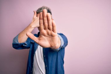 Pembe izole edilmiş arka planda gözlük ve kot ceket giyen genç, yakışıklı, modern bir adam ellerle gözleri kaplıyor ve üzgün ve korku ifadeleriyle dur hareketi yapıyor. Utanç verici ve olumsuz bir kavram..
