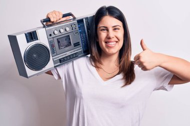 Genç ve güzel bir kadın beyaz arka planda klasik müzik çalar kullanarak müzik dinliyor mutlu ve pozitif gülümsüyor, başparmağıyla mükemmel ve onay işareti yapıyor.