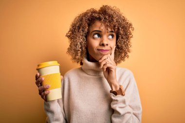 Genç Afro-Amerikalı kıvırcık kadın sarı arka planda bir fincan kahve içiyor. Ciddi bir yüz ifadesi var. Soru sormayı düşünüyor.