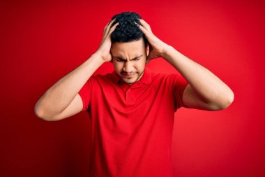 Kırmızı polo giyen yakışıklı genç adam izole edilmiş arka planda dikiliyor. Acı ve migren yüzünden çaresiz ve stresli bir baş ağrısı çekiyor. Ellerini başının üstüne koy..