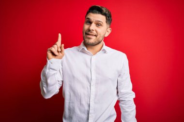 Mavi gözlü, zarif gömlekli, kırmızı izole edilmiş arka planda duran genç bir iş adamı başarılı bir fikri işaret ediyor. Heyecanlı ve mutlu. Bir numara..