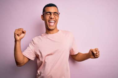 Yakışıklı Afro-Amerikan erkeği pembe arka planda günlük tişört ve gözlük takıyor mutlu ve neşeli dans ediyor, hareketli, rahat ve kendinden emin bir şekilde müzik dinliyor.