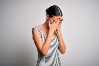Genç, güzel esmer kadın, izole edilmiş beyaz arka planda günlük çizgili elbiseler giyiyor ve ağlarken suratını ellerle kapatıyor. Depresyon kavramı.