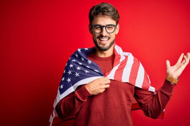 Gözlüklü genç adam ve Amerika Birleşik Devletleri bayrağı izole edilmiş kırmızı arka planda çok mutlu ve heyecanlı, kazanan yüz ifadesi zaferi büyük bir gülümsemeyle kutluyor ve ellerini kaldırıyor.