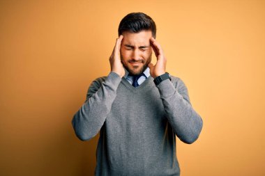 Yakışıklı genç işadamı sarı arka plan üzerine zarif bir kazak ve kravat giyip acı ve migren yüzünden çaresiz ve stresli bir baş ağrısı çekiyor. Ellerini başının üstüne koy..