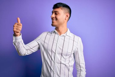 Zarif iş gömleği giyen genç yakışıklı İspanyol adam mor arka planda duruyor gururlu görünüyor, gülümsüyor yan tarafa baş parmak işareti yapıyor.