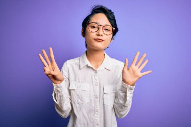 Genç, güzel Asyalı bir kız. Günlük gömlek ve gözlük takıyor. Mor arka planda duruyor..