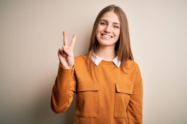 Genç, güzel, kızıl saçlı bir kadın izole edilmiş beyaz bir arka plan üzerine günlük süveter giyiyor ve iki numaralı parmaklarıyla işaret ederken kendinden emin ve mutlu gülümsüyor..
