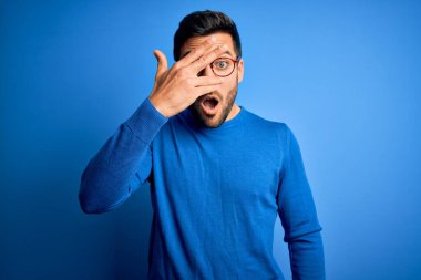 Sakallı, sıradan süveter giyen ve mavi arka planda gözlüklü genç yakışıklı bir adam. Şok içinde yüzünü ve gözlerini kapayarak utangaç bir ifadeyle parmaklarının arasından bakıyor..