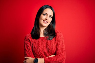 Mavi gözlü genç esmer kadın izole edilmiş kırmızı arka planda günlük süveter giyiyor ve kameraya bakarak kollarını kavuşturup gülümsüyor. Pozitif kişi.