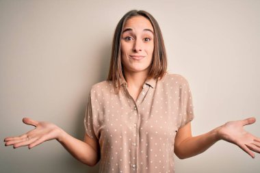 İzole edilmiş beyaz arka plan üzerinde duran sıradan bir gömlek giyen genç, güzel bir kadın. Kolları ve elleri havada, kafası karışmış bir ifade. Şüphe kavramı.