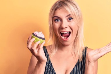 Sarışın, güzel bir kadın, izole edilmiş sarı arka planda tatlı çikolatalı kek yerken mutluluğu ve mutlu bir gülümsemeyle kazanan ifadeyi el kaldırarak kutluyor.