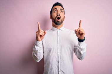 Genç, sakallı, rahat gömlekli, pembe arka planda dikilen yakışıklı adam şaşırmış ve şaşırmış bir şekilde yukarı bakıp parmaklarını göstermiş ve kollarını kaldırmış..