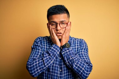 Sarı arka plan üzerine gündelik gömlek ve gözlük takan genç, yakışıklı latin adam. Yüzü, depresyonu ve üzüntüyü örten yorgun eller, sorun yüzünden üzgün ve tedirgin.