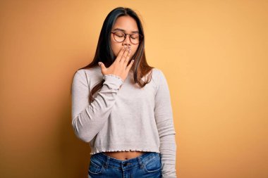 Sarı arka planda günlük süveter ve gözlük takan genç, güzel Asyalı kız ağzıyla ağzını kapatmaktan sıkılmış. Huzursuz ve uykulu..