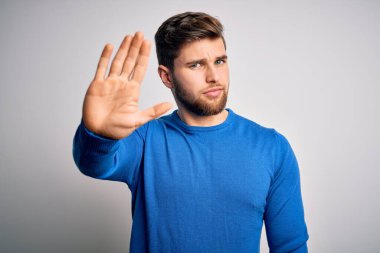 Young handsome blond man with beard and blue eyes wearing casual sweater doing stop sing with palm of the hand. Warning expression with negative and serious gesture on the face.