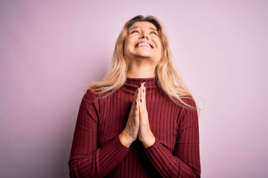 İzole edilmiş pembe arka plan üzerine gündelik süveter giyen genç, güzel sarışın bir kadın elleri bağlı yalvarıyor ve dua ediyor. Yüzünde umut ifadesi var. Çok duygusal ve endişeli. Yalvarıyorum..