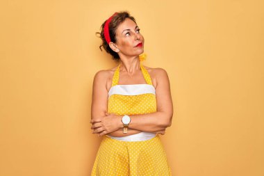 50 'li yaşlarda, sarı arka plan üzerine eski tarz elbise giyen, elleri çenesinde soru soran, düşünceli ifadeli orta yaşlı bir kadın. Düşünceli bir yüzle gülümsüyor. Şüphe kavramı.