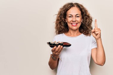 Orta yaşlı güzel bir kadın, elinde maki suşi tepsisiyle izole edilmiş beyaz arka planda bir fikir ya da soru işareti ile gülümsüyor.