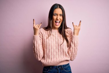 Genç, güzel esmer bir kadın pembe arka plan üzerine günlük süveter giyip çılgın bir ifadeyle elleri havada rock sembolü yaparak bağırıyor. Müzik yıldızı. Ağır müzik konsepti.