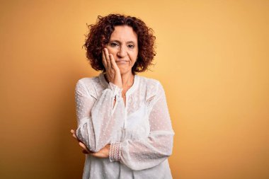 Orta yaşlı güzel kıvırcık saçlı kadın sarı arka planda rahat bir yaz elbisesi giyiyor. Kollarını kavuşturup depresyondan sıkılmış ve yorgun görünüyor..