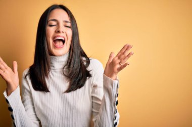 Mavi gözlü genç esmer kadın izole edilmiş sarı arka plan üzerine gündelik süveter giyerek başarının çılgınlığını kollarını kaldırarak ve gözlerini kapayarak heyecanla bağırarak kutluyor. Kazanan konsept