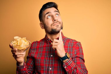 Elinde patates cipsi olan genç yakışıklı adam izole edilmiş sarı arka planda kendini işaret eden sürpriz yüzlü.