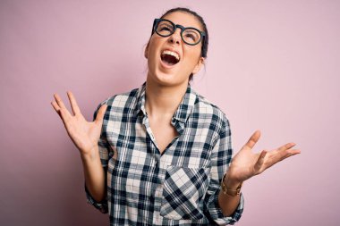 Genç, güzel, esmer bir kadın pembe arka plan üzerine günlük gömlek ve gözlük takıyor deli deli deli bağırıyor ve agresif bir ifade ve kollar havada bağırıyor. Hayal kırıklığı konsepti.