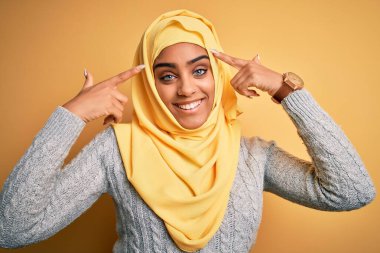 Genç ve güzel Afro-Amerikan kızı, izole edilmiş sarı arka plan üzerinde Müslüman başörtüsü takıyor. İki eliyle başını gösteren gülümseme, harika bir fikir ya da düşünce, iyi bir hafıza.