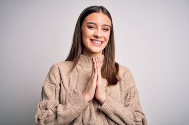 Young beautiful brunette woman wearing casual sweater standing over white background praying with hands together asking for forgiveness smiling confident.