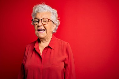 Son sınıf güzeli, gri saçlı, rahat gömlekli, kırmızı arka planda gözlüklü seksi bir ifadeyle kameraya bakan, neşeli ve mutlu yüzlü bir kadın..