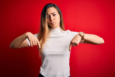 Mavi gözlü güzel sarışın kadın kırmızı arka planda günlük beyaz tişört giyiyor üzgün ve üzgün görünüyor, parmaklarıyla yön gösteriyor, mutsuz ve depresif..