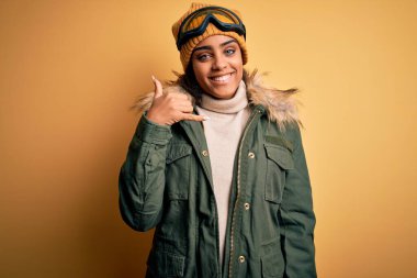 Genç Afrikalı Afro-Amerikan kayakçı kız kar sporu kıyafetleri ve kayak gözlüğü takıp gülümsüyor telefonla el ve parmaklarıyla telefonla konuşuyor gibi yapıyor. İletişim kavramları.