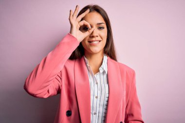 Güzel, esmer, ceketli bir kadın pembe arka planda duruyor. El hareketleriyle gülümsüyor ve mutlu yüzüyle parmaklarının arasından bakıyor..