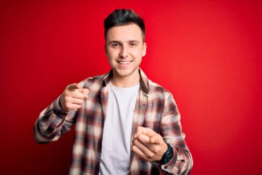 Kırmızı izole arka plan üzerine gündelik modern tişört giyen yakışıklı beyaz adam mutlu ve komik yüzlü parmaklarını kameraya doğrultuyor. İyi enerji ve titreşimler.