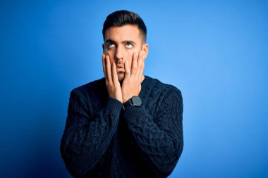 İzole edilmiş mavi arka plan üzerinde duran genç, yakışıklı bir adam. Yüzü, depresyonu ve üzüntüyü örten yorgun eller, sorun yüzünden üzgün ve sinirli.