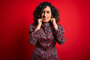 Genç ve güzel kıvırcık Arap kadın gündelik çiçekli elbiseler giyiyor kırmızı arka planda dikiliyor yüksek sesli müzik sesi için parmaklarıyla kulaklarını kapatıyor. Sağır kavramı.