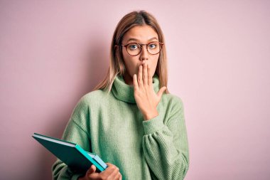 Gözlük takan genç bir kadın pembe arka plan örtüsünün üzerine kitap tutuyor. Ağzı ve elleri hata yüzünden şoke olmuş, korkunun ifadesi, sessizlikten korkmak, gizli konsept.