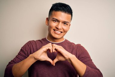 İzole edilmiş beyaz arka plan üzerinde dikilmiş, elleri ile kalp sembolü ve şekli gösteren bir gülümsemeyle genç, yakışıklı Latin bir adam. Romantik konsept.