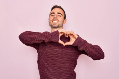 Mavi gözlü, sıradan bir süveter giyen, pembe arka planda duran, aşık olmuş, elleriyle kalp sembolü yapan genç yakışıklı bir adam. Romantik konsept.